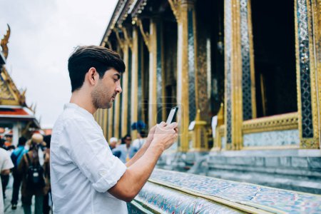 Photo for Pensive spanish man tourist using app on mobile phone making photo share in blog visiting asian city during trip, millennial spanish guy shooting video on smartphone  standing on crowded square - Royalty Free Image