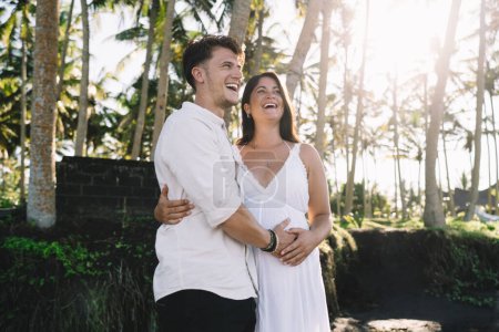 Téléchargez les photos : Delightedromantic femme enceinte en robe d'été blanche collant avec mari heureux et toucher le ventre tout en profitant week-end dans un parc tropical ensoleillé - en image libre de droit