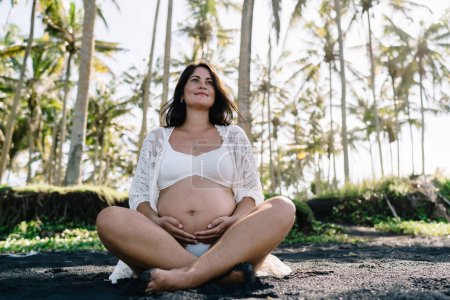 From below of full body happy pregnant female in bikini and beach cover up with crossed legs sitting on wet ground and touching belly against tropical trees in forest