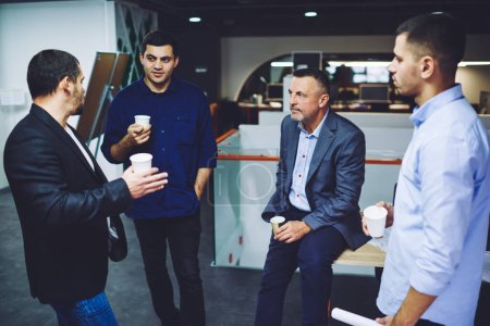 Group of confident male employees in smart casual clothing discussing collaborative work and trade exchange, businessmen with takeaway coffee to go communicate about financial marketing indoors