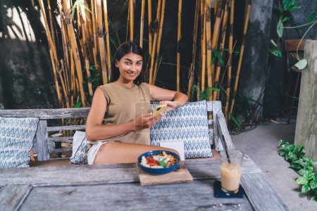 Foto de Mujer étnica positiva en ropa casual sonriendo y mirando a la cámara mientras se relaja en un cómodo banco de café tropical con teléfono inteligente en las manos - Imagen libre de derechos