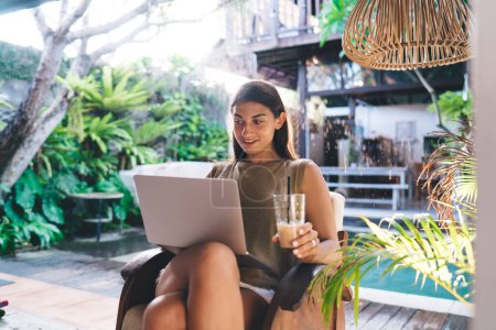 Téléchargez les photos : Jeune contenu ethnique femme employée à distance avec netbook et verre de boisson rafraîchissante assis avec les jambes croisées dans le fauteuil au bord de la piscine et regardant loin - en image libre de droit
