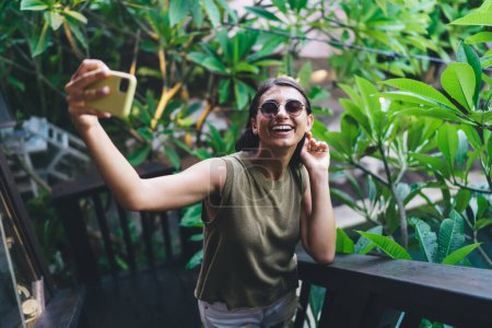 Téléchargez les photos : Angle élevé de femme heureuse en tenue décontractée et lunettes de soleil souriant et prenant selfie sur téléphone mobile pendant les vacances d'été sur Bali - en image libre de droit