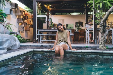 Téléchargez les photos : Jeune voyageuse ethnique souriante portant des vêtements d'été assis près de la piscine avec de l'eau bleue et profitant de vacances dans un pays chaud exotique - en image libre de droit