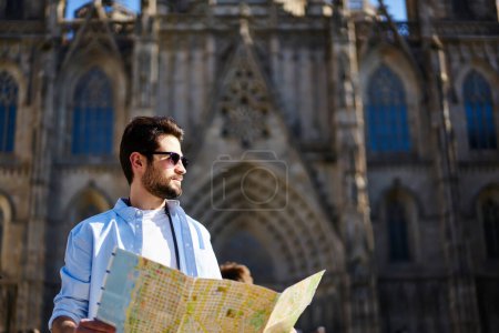 Photo for Low angle of pensive bearded male traveler in casual outfit and sunglasses looking away while holding map in hands during walk - Royalty Free Image