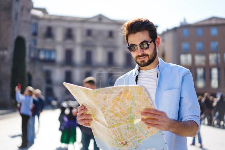 Zufriedene bärtige männliche Touristen in lässigem Outfit und Sonnenbrille lesen Karte, während die Wahl der Lage auf der Straße während des sonnigen Sommertages