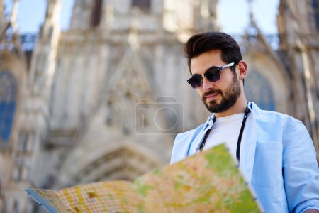 Low angle of serious bearded male traveler in casual outfit and sunglasses navigating with map while walking on street during sightseeing