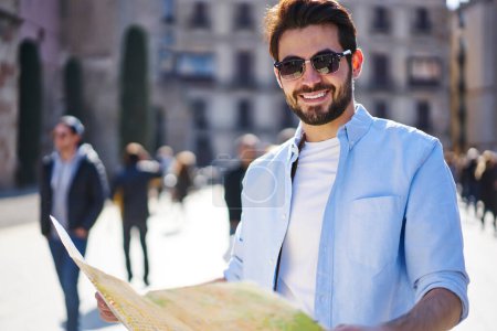 Cheerful bearded man in trendy outfit and sunglasses looking at camera and smiling while navigating on street with map during walk