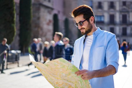 Positiver bärtiger Mann in lässiger Kleidung und Sonnenbrille, der bei der Routenanalyse die Landkarte betrachtet und bei sonnigem Sommerurlaub lächelt