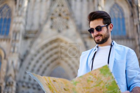 From below of cheerful bearded male traveler in casual wear and sunglasses standing against old building with map and smiling