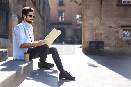 Foto de Vista lateral de cuerpo completo de un joven viajero barbudo con ropa casual y gafas de sol mirando hacia otro lado mientras descansa en las escaleras a la luz del sol y leyendo el mapa - Imagen libre de derechos