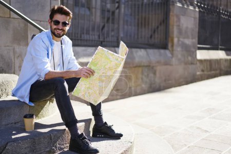 Volle Länge von gut gelaunten männlichen Touristen in Sonnenbrille mit Bart, die auf Treppen lümmeln und mit hellem zahmen Lächeln eine große Stadtkarte aus Papier lesen.