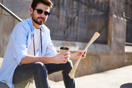 Male traveler with beard in light blue shirt and sunglasses drinking beverage and reading map of city while resting in yard of historical building