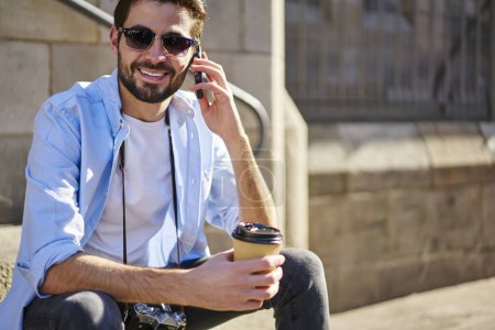 Foto de Alegre turista masculino en ropa casual con cámara fotográfica hablando en el teléfono móvil con sonrisa brillante mientras descansa en el patio y beber café - Imagen libre de derechos