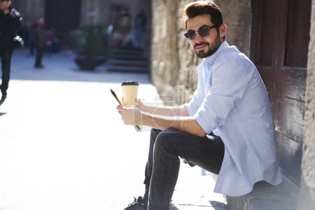 Téléchargez les photos : Jeune touriste masculin en chemise bleu clair et jeans avec barbe assis sur le trottoir et boire du café tout en se prélassant dans la cour du bâtiment historique - en image libre de droit