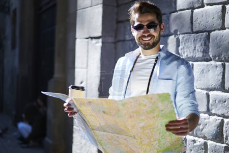 Cheerful bearded man in casual outfit and sunglasses holding paper road map and cup of coffee to go while standing near stone building