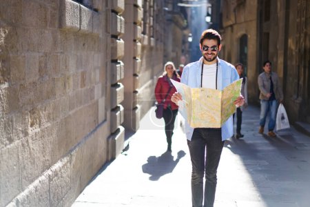 Smiling young male tourist in casual outfit and sunglasses strolling on sidewalk between old buildings and exploring tourist routes on paper map