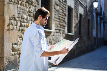 Photo for Side view of focused ethnic male in casual clothes and sunglasses walking on street near shabby building and examining location of town with paper map on sunny day - Royalty Free Image