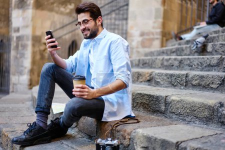 Téléchargez les photos : Vue latérale pleine longueur du jeune homme ethnique souriant en tenue décontractée et lunettes avec café à emporter et appareil photo à proximité à l'aide d'un smartphone tout en étant assis sur des escaliers en pierre du vieil immeuble - en image libre de droit