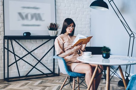 Kaukasisches Hipstermädchen in stylischer Brille zur Sehkorrektur liest während des Freizeit-Wochenendes in einer modernen Wohnung interessante Literaturbücher, junge Frau interessiert sich für Buchhandlung, während sie zu Hause ausruht