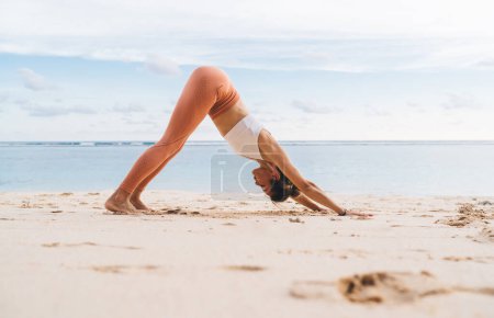 Téléchargez les photos : Vue latérale de la dame mince en vêtements de sport étirant le corps vers le bas face à la pose de chien sur le sable de la côte exotique tout en méditant seul tôt le matin - en image libre de droit