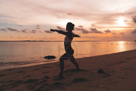 Téléchargez les photos : Silhouette de femme anonyme en tenue de sport pratiquant Virabhadrasana II asana tout en faisant du yoga sur une plage de sable près de l'océan calme au coucher du soleil - en image libre de droit
