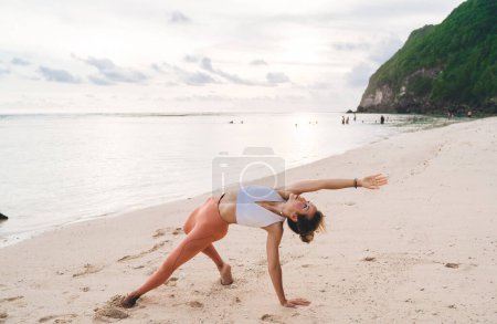 Téléchargez les photos : Plage de sable propre près des montagnes verdoyantes et jeune femme en legging orange pratiquant Fallen Triangle asana et regardant le ciel - en image libre de droit