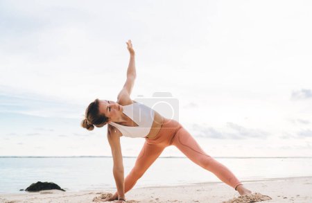 Téléchargez les photos : Corps complet de sportive flexible concentrée debout sur la plage de sable près de la mer calme et faire du yoga asana seul pendant la journée - en image libre de droit