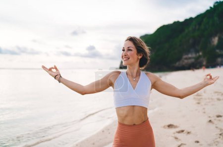 Téléchargez les photos : Jeune femme active en vêtements de sport légers debout sur la côte près de la mer et regardant loin tout en montrant mudra gyan avec les mains dans la journée ensoleillée - en image libre de droit