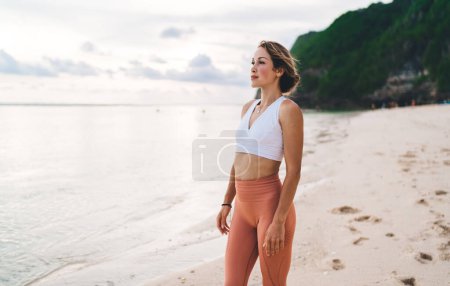 Foto de Ajuste mujer de buen aspecto en ropa deportiva admirando la belleza del océano tranquilo mientras disfruta de vacaciones en la playa en el magnífico exótico Bali - Imagen libre de derechos