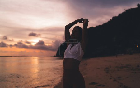 Téléchargez les photos : Femme mince debout seul sur le bord de la mer et profiter de la liberté tout en faisant des exercices de respiration contre l'océan calme et ciel nuageux coucher de soleil - en image libre de droit