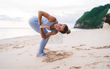 Foto de Cuerpo completo descalzo alegre hembra en ropa deportiva de pie en la playa de arena y la práctica de la silla giratoria posan en Bali mientras hace yoga - Imagen libre de derechos