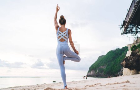 Téléchargez les photos : Vue de dos de yogi femelle mince debout à Tree Pose et méditation pendant l'entraînement matinal à la plage, femme athlétique avec une forme corporelle parfaite relaxante pendant la pratique sportive au bord de la mer - en image libre de droit