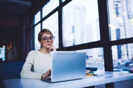 Téléchargez les photos : Angle bas de freelance féminine heureuse avec des lunettes regardant la caméra assise à la table près de la fenêtre de navigation ordinateur portable tout en travaillant - en image libre de droit