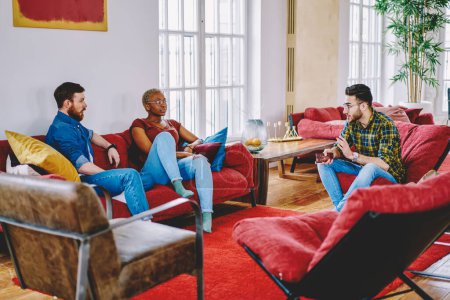 Diverse beste Freunde in Freizeitkleidung kommunizieren miteinander, ruhen sich an bequemen Möbeln in einer modernen Wohnung aus, junge Hipster unterhalten sich drinnen und verbringen ihre Freizeit gemeinsam zu Hause.