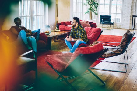 Männer und Frauen diskutieren über Freundschaft, während sie auf roten Möbeln im Wohnbereich ruhen, multiethnische Hipster in lässigem Outfit kommunizieren über modernes Design in einer flachen Wohnung