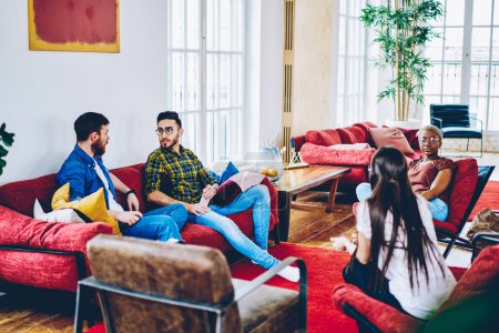 Young male and female guests discussing friendship relations during weekend meeting in loft apartment with comfortable red furniture, students talking about design project during leisure time