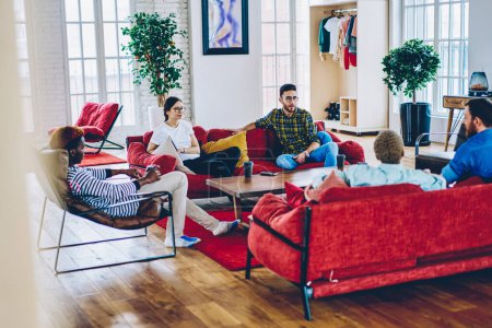 Jugendliche 20er Jahre sitzen auf der Couch im Wohnzimmer und unterhalten sich gastfreundlich mit den Gästen, multiethnische Freunde kommunizieren beim Kaffeetreffen zu Hause im trendigen Loft-Design