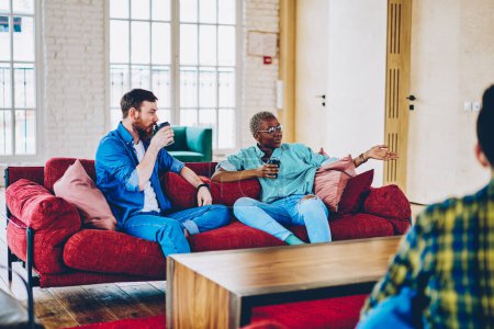 Gruppe multikultureller Menschen in Freizeitkleidung sitzt auf Sofa mit Koffeingetränk in moderner Wohnung, diverse Hipster genießen freundliches Treffen zum Ausruhen und Kaffeetrinken, um nach drinnen zu gehen