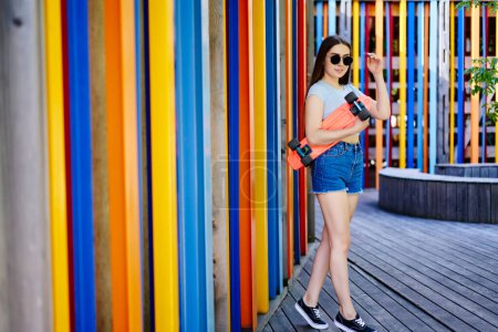 Foto de Retrato de larga duración de una chica hipster milenaria vestida con ropa casual sosteniendo un tablero de peniques y mirando a la cámara durante el día de verano para recrear a un joven patinador adolescente con gafas de sol - Imagen libre de derechos