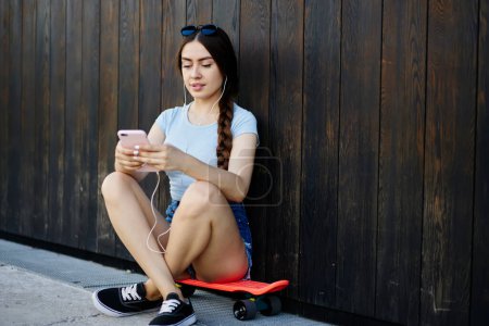 Téléchargez les photos : Attrayant hipster girl dans les écouteurs navigation contenu multimédia dans les réseaux sociaux e en utilisant le téléphone mobile pendant les loisirs à la rue, millénaire utilisateur écoutant de la musique playlist de smartphone gadget - en image libre de droit