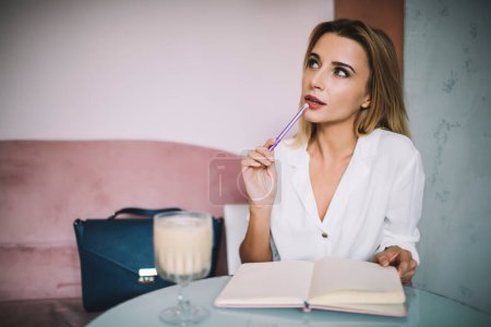 Mujer joven soñadora en blusa blanca sentada en mesa redonda con un vaso de café y mirando hacia otro lado tocando cuidadosamente el labio con pluma