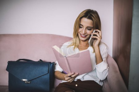 Téléchargez les photos : Jeune femme souriante en chemisier blanc lisant des notes dans un carnet ouvert alors qu'elle était assise sur un canapé près d'un sac et parlait au téléphone - en image libre de droit