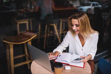 Foto de Desde arriba de la mujer de negocios con el teléfono móvil y el cuaderno de escribir informe en netbook en la mesa en la cafetería de la calle en el día - Imagen libre de derechos