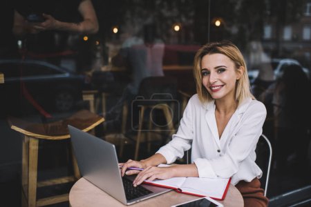 Téléchargez les photos : Entrepreneur féminin positif avec des cheveux blonds dans des vêtements élégants assis à table avec ordinateur portable et netbook de navigation tout en travaillant sur la terrasse du café en journée - en image libre de droit