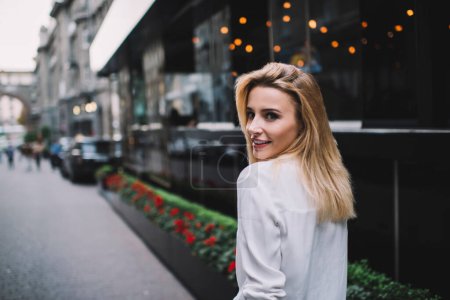 Rückansicht einer fröhlichen jungen blonden Frau in weißer Bluse, die durch ein modernes Gebäude auf der Straße schlendert und über die Schulter schaut