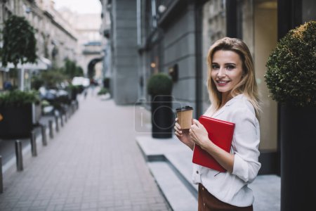 Foto de Joven estudiante alegre en blusa blanca con cuaderno mirando a la cámara mientras camina por la calle y bebe café para ir - Imagen libre de derechos
