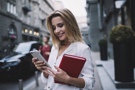 Téléchargez les photos : Femme entrepreneur élégante avec des cheveux blonds en vêtements formels debout sur la rue et smartphone de navigation tout en marchant le long du centre-ville - en image libre de droit