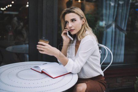 Téléchargez les photos : Vue latérale de la femme aux cheveux blonds graves dans des vêtements élégants ayant une conversation sur smartphone tout en buvant du café à emporter dans un café extérieur - en image libre de droit