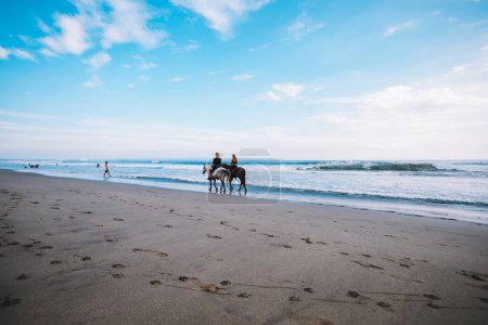 Back view of distant unrecognizable female friends riding horses near wavy sea on sandy seaside and enjoying sunny summer day